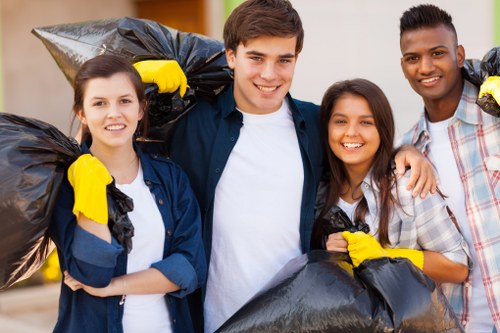 Four young adults, two women and two men, standing outdoors on a paved surface with a blurred background of trees and residential buildings. They are smiling and dressed casually, each wearing yellow gloves, and holding large black rubbish bags filled with waste, indicating participation in a rubbish removal or cleanup activity for garden or household waste. The individuals are positioned closely together, suggesting camaraderie and teamwork, with some bags resting on their shoulders. The scene appears to take place during daytime with natural lighting, reflecting an active effort to clear rubbish in a community setting. This imagery aligns with professional waste management services offered by companies like Garden Clearance Deptford in the area of Deptford, South-East London, focusing on rubbish clearance for gardens or properties.