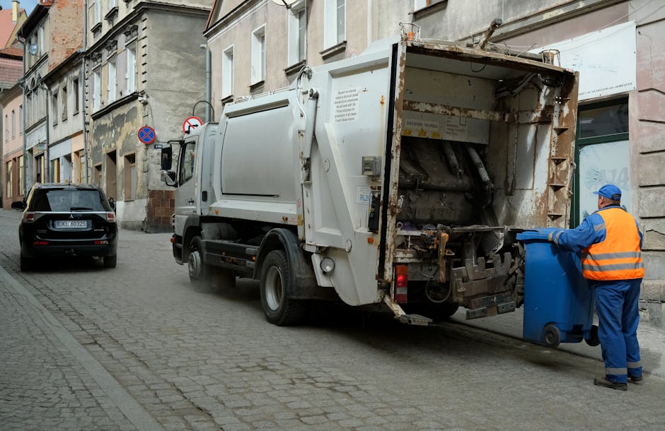 A street scene showing a waste collection worker in a high-visibility vest operating a large red rubbish truck. The worker is standing at the rear of the truck, which is positioned on the side of the 