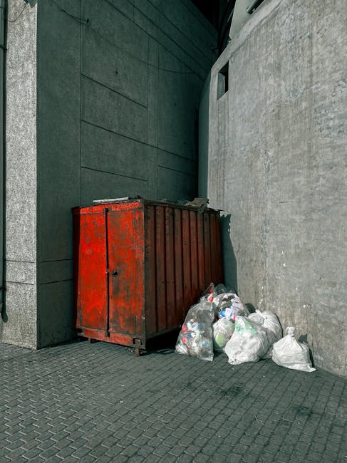 A collection of several black rubbish bags piled against the green-painted exterior wall of a building, situated next to a set of wooden stairs leading up to a door with a small porch. The bags appear