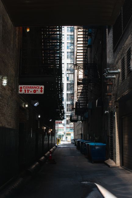 A city street scene featuring a variety of multi-story buildings with diverse architectural styles, including brick facades, large rectangular windows, and decorative elements. In the foreground, ther